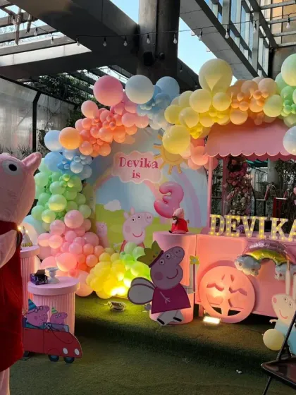 The Peppa Pig mascot stands proudly next to the custom-designed backdrop, ready to welcome guests to Devika's 3rd birthday party.