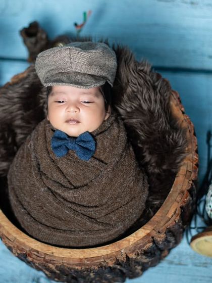 A dapper little newborn in a flat cap and bow tie, swaddled and sleeping in a wooden bowl.