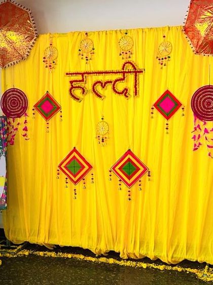 An indoor Haldi and Mehendi corner, personalized with a custom welcome easel board. The backdrop features a traditional yellow curtain with decorative umbrellas and hangings, creating a dedicated space for the ceremonies.