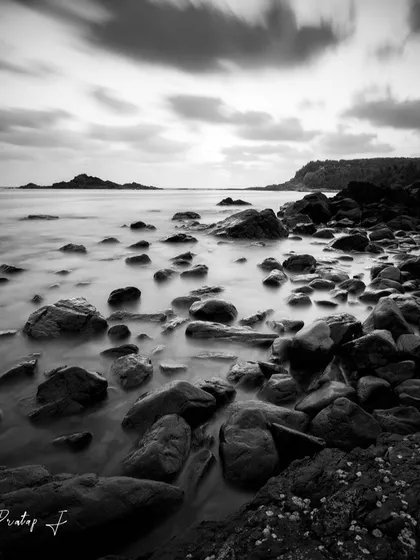 A long exposure of a rocky shoreline in Gokarna, rendered in black and white. The motion-blurred clouds and water contrast with the sharp, barnacle-covered rocks.