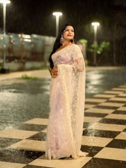 A full-length elegant pose. The checkerboard pattern of the ground contrasts beautifully with the soft, flowing saree.