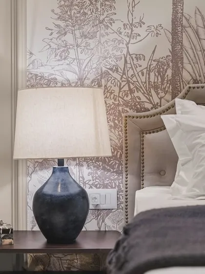 A detailed shot of the bedside table, focusing on the textures and tones of the space. The dark blue ceramic lamp provides a beautiful contrast against the soft, monochrome wallpaper and plush bedding.