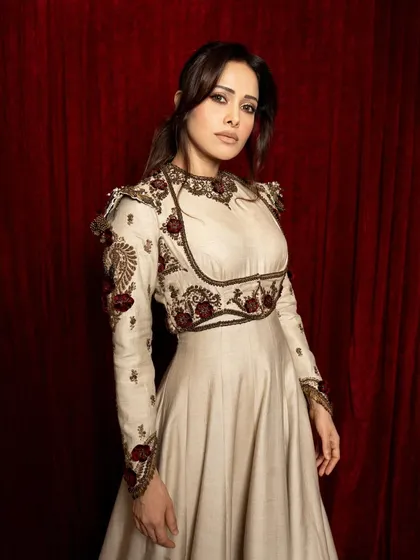 A portrait of Nushrratt Bharuccha with an 'Umrao Jaan' inspired theme. The rich red curtain and traditional outfit are lit to create a regal, cinematic feel.