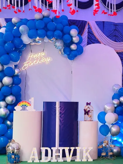 A wider view of the Adhvik's birthday decoration, showing the full arrangement of the balloon arch, backdrop, and props like the decorative bird cage and rainbow sign.