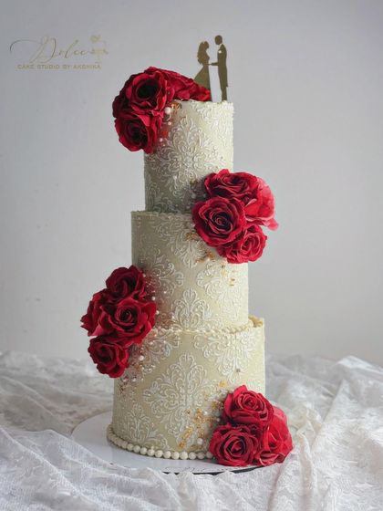 This grand, three-tiered cake features an intricate white stencil pattern over an ivory base, adorned with clusters of rich, red sugar roses. A timeless design for a wedding or grand anniversary.