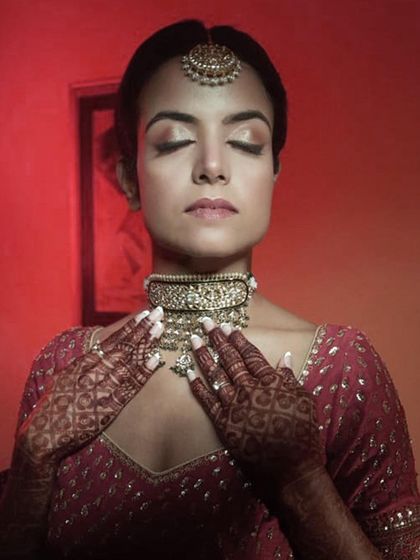 A tranquil portrait of a bride with her eyes closed, showcasing the soft, blended eyeshadow and flawless skin finish. Her henna-adorned hands gently frame her face.
