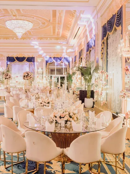 A regal welcome dinner setup in a grand ballroom, with opulent gold and blue decor, plush white seating, and stunning floral centerpieces.