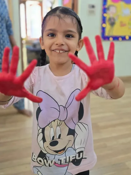 Getting a little messy is part of the fun and learning process here. This happy smile, framed by red-painted hands, captures the pure delight of our sensory art activities that help develop fine motor skills.
