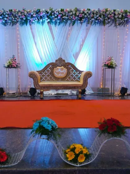 This reception decor features a wide stage with an elegant gold-carved sofa. The backdrop is made of pleated white drapes with blue uplighting, fairy lights, and a full floral border across the top, creating a dreamy and sophisticated look.