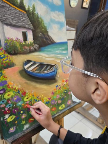 A close-up of the young artist's hands as he adds colorful flowers to the foreground of his oil pastel landscape.