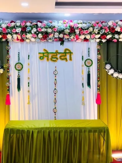 This Mehandi backdrop uses a combination of white and green drapes with a top border of colorful flowers. The hanging floral rings and tassels add a delicate and festive touch.