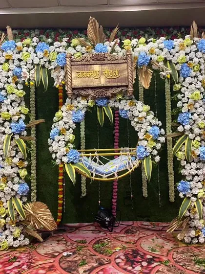 A luxurious floral naming ceremony (Janam Sohala) stage. The baby's cradle is framed by dense arrangements of artificial flowers in blue, white, and yellow, set against a green wall for a rich, traditional feel.