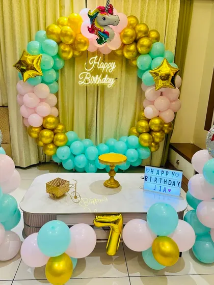 A unicorn themed birthday decoration for a 7-year-old's home party. This setup includes a full ring of pastel and gold balloons, a unicorn foil balloon, and a light box with a personalized birthday message.