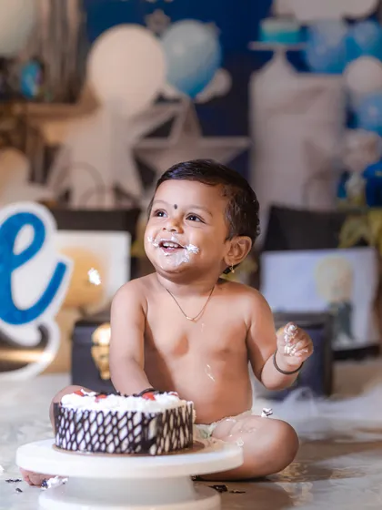 A happy baby boy with cake on his face, looking up with a huge smile during his blue-and-white themed cake smash session.