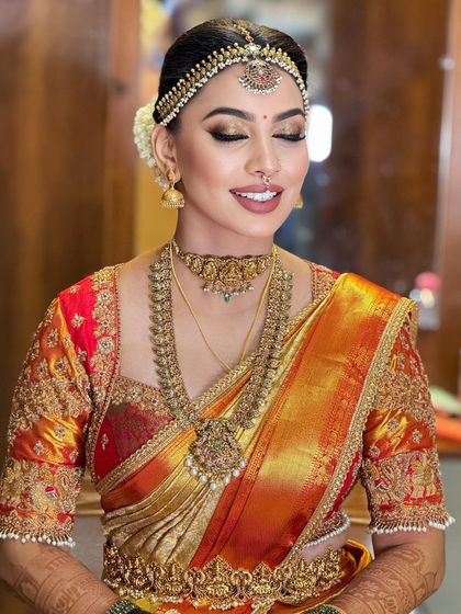 A happy bride in a vibrant orange and red Kanjeevaram. The makeup is warm and glowing, with a focus on golden highlights and a nude lip.