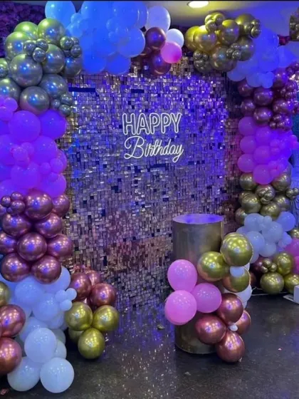 For a glamorous first birthday, I can create a stunning photo corner with a silver shimmer wall. This setup is framed by balloon columns in purple, pink, and metallic tones, and includes a 'Happy Birthday' neon sign and a light-up number '1'.