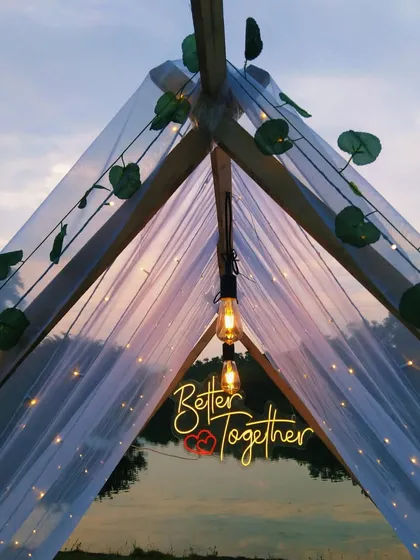 A romantic and intimate anniversary celebration setup in a tent by a lake in Karjat. The warm glow of the 'Better Together' neon sign and fairy lights creates a cozy, unforgettable atmosphere at dusk.