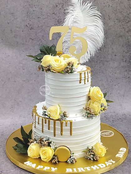 A grand two-tiered cake for a 75th birthday. This elegant design features a golden drip, yellow roses, and a large feather for a touch of glamour.