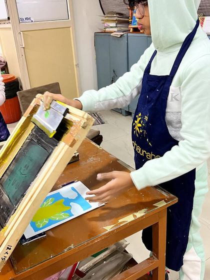 A young student learning the process of serigraphy, or silkscreen printing. He's carefully checking his multi-color print of a cute character.