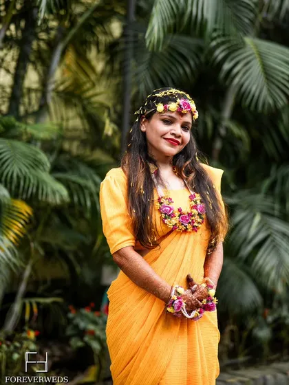 A beautiful portrait of the Haldi bride, Nisha, adorned with floral jewelry. The lush green background provides a stunning contrast to her yellow saree, making the colors pop.