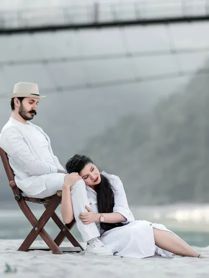 A pensive and artistic shot of the couple on the riverbank, with the bridge faintly visible in the background. The mood is calm and romantic.