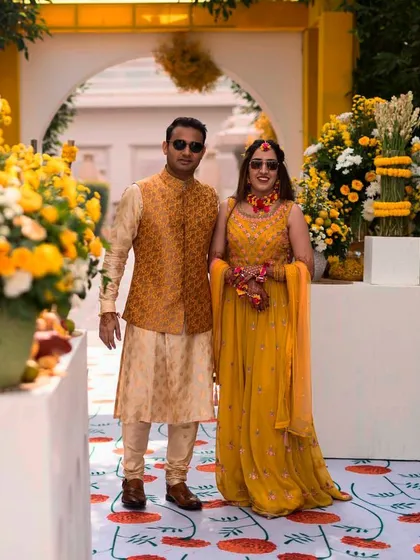 The couple posing at their Haldi ceremony. The decor features a mix of yellow and white flowers, with a custom-designed carpet that adds a modern, graphic element.