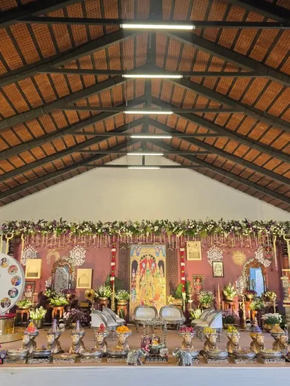 A wide-angle view of a traditional naming ceremony (Namakarna) I decorated. The setup includes a custom backdrop with a deity portrait, a silver cradle for the baby, and an extensive arrangement of silver cow props holding offerings, all under a high, wood-beamed ceiling.