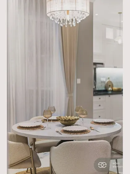A close-up of the elegantly set dining table, with warm-toned glassware and a central brass bowl. The soft lighting from the chandelier and the gentle folds of the curtains create a warm and inviting atmosphere.
