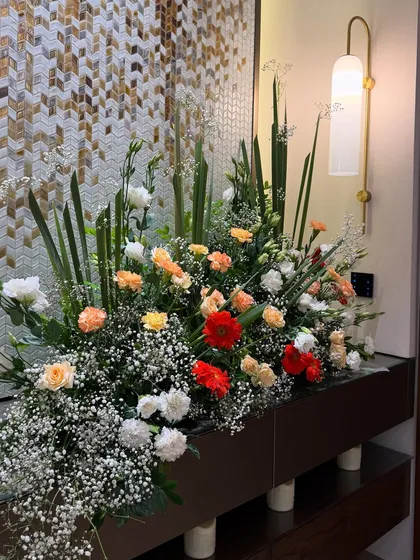 A bountiful floral arrangement designed to fill a console space. The mix of red, orange, and white flowers with baby's breath adds a vibrant pop of color against the home's modern, textured wall.