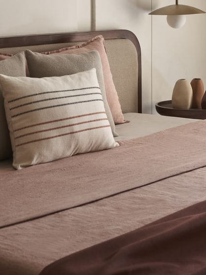 The layers of linen bedding in dusty rose and terracotta tones add warmth and comfort. The accent cushion with simple stripes provides a touch of handcrafted detail.