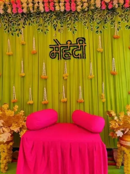 A simple and sweet Mehandi seating with a lime green backdrop and hanging floral buds. The bright pink gadda and golden flower pots create a vibrant and cheerful setting.