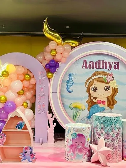 We dove deep to create this magical mermaid world for Aadhya's first birthday. This elaborate stage includes a custom backdrop with a porthole view, mermaid-scale patterns, a boat-shaped shelf for favors, and a light-up number '1'.