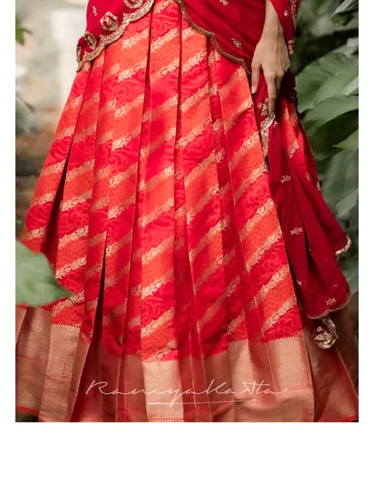 A detail shot of the orange-red brocade lehenga skirt, highlighting the rich texture and golden weaving of the traditional fabric.