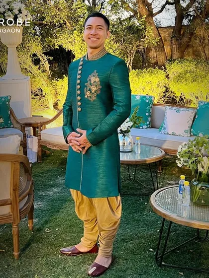 This happy customer looks sharp in a teal green silk Indo-western sherwani with asymmetrical gold embroidery. It's a unique and vibrant choice for a Mehendi or Sangeet ceremony.