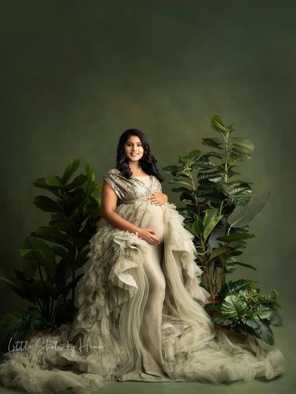 A beautiful, nature-themed portrait of an expecting mother. She is seated in a stunning silvery-green gown amidst large tropical plants against a matching green backdrop.