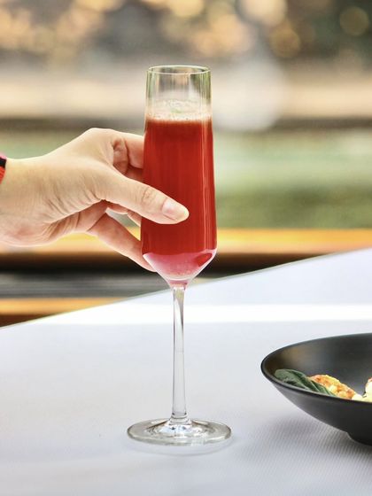 A guest enjoys one of our vibrant cocktails at a table overlooking the water. This image captures the essence of the Indian Accent experience, where every detail, from the drink in your glass to the view, comes together beautifully.