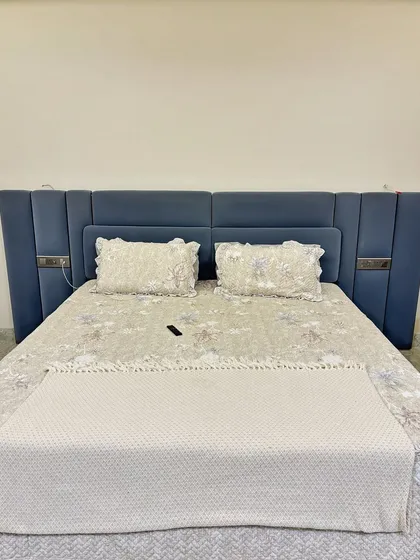 This custom bed features an extra-wide upholstered headboard in a calming blue fabric. The side panels include sleek metallic accents and integrated switches for a clean, modern look.