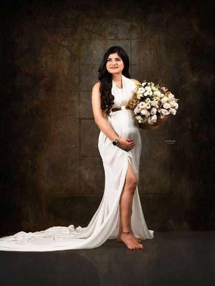 A beautiful fine art portrait against a dark, classic backdrop. The client looks serene in a simple white gown, holding a bouquet of flowers, creating a timeless and elegant image.