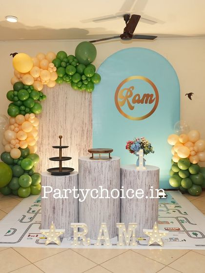 A simple yet elegant home decoration for a first birthday. We used a pastel blue arched backdrop, wood-textured pedestals, and a green and peach balloon arrangement for a soft, modern look.