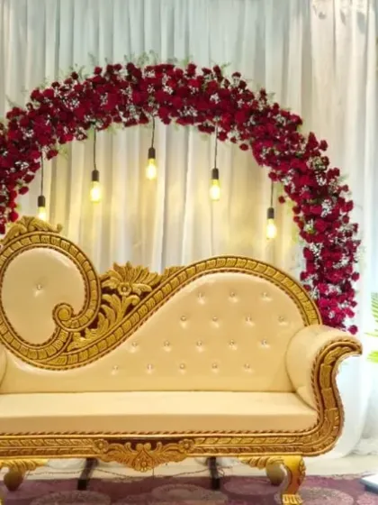 An elegant stage for a baby shower function. The decor features a circular arch of deep red flowers against a white drape, with a classic cream-colored diwan for seating.