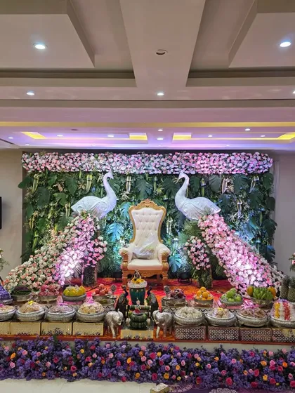 A unique and vibrant stage for a half-saree ceremony, featuring two large peacock figures made of flowers. The backdrop is a lush wall of green leaves, creating a beautiful contrast with the pink and white floral peacocks and the ornate central throne.