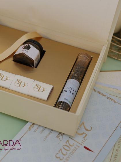 A modern and minimalist gift box in a neutral cream color. It contains a curated selection of treats, including a jar of gourmet spread, a test tube of tea or spices, and monogrammed chocolates.