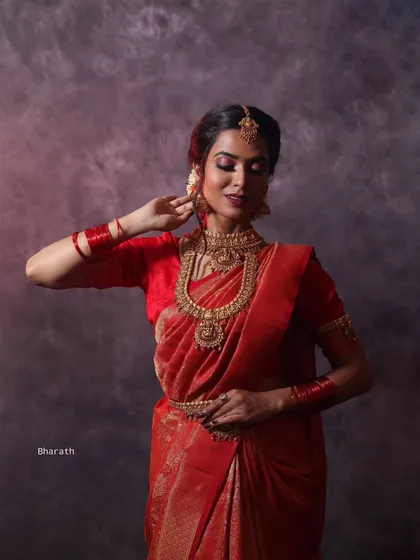 A dramatic, low-light studio portrait in a red bridal saree. The lighting creates a spotlight effect, focusing entirely on the model and the richness of her traditional attire.