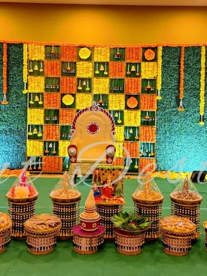 This is a complete setup for a half-saree function, showing both the main stage and the traditional seer plate arrangement. The backdrop features a unique checkerboard pattern of fresh marigolds and green leaves.