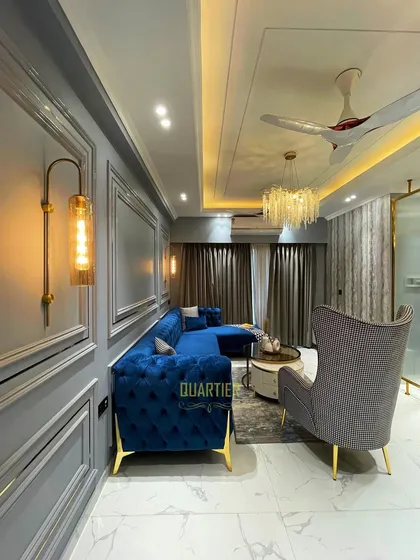 A shot of the living room at Elite Golf Greens, inspired by European design. The space features grey walls with trim moulding, a luxurious blue velvet sofa, and gold accents on the furniture and lighting.