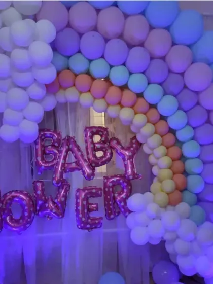 A beautiful rainbow balloon arch against a sheer curtain backdrop for a home baby shower. The 'Baby Shower' foil balloons in pink add a celebratory touch to this colorful and happy setup.
