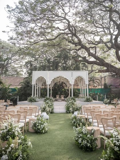 The main wedding mandap, a grand white structure with multiple arches, set under a majestic old tree, blending architectural design with nature.