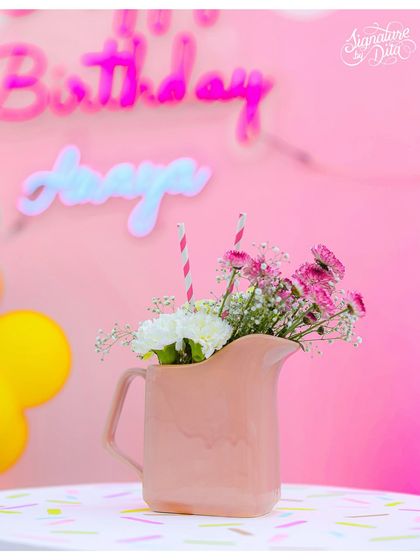 A simple, sweet detail. A pink pitcher holding fresh flowers with a 'Happy Birthday' neon sign glowing in the background.