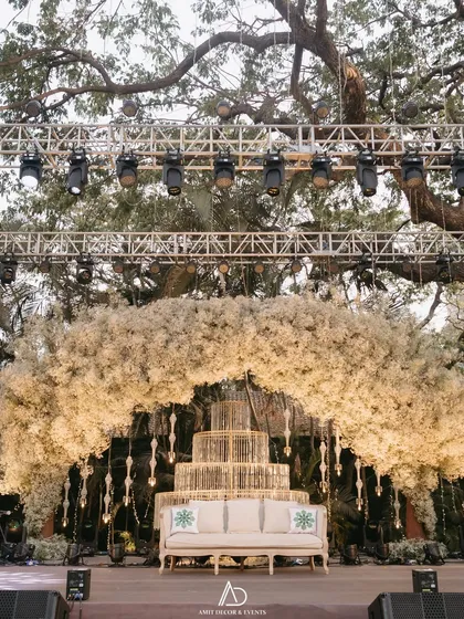 The main stage for an outdoor engagement, featuring a stunning arch of white baby's breath and a multi-tiered cake display. The entire setup is framed by a magnificent old tree and a professional lighting rig.