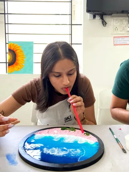 A participant uses a straw to create delicate wave effects in her pink-hued resin beach art.